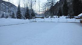 Ein schneebedeckter Platz umgeben von Bäumen und Bergen. Die Szene strahlt eine ruhige Winteratmosphäre aus.