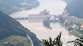 Donaukraftwerk-Jochenstein in Engelhartszell: Eine bergige Landschaft mit einem großen Gebäudekomplex an einem See. Die Architektur des Gebäudes wirkt traditionell und historisch. Die Umgebung ist von dichten Wäldern und grünen Hügeln geprägt. Insgesamt vermittelt das Bild eine idyllische und malerische Szenerie.