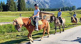 Reiterinnen auf Pferden genießen einen sonnigen Tag in den Bergen. Die Umgebung ist grün, umgeben von majestätischen Bergen.