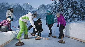 Eine Gruppe von Menschen spielt Eisstockschießen auf einer zugefrorenen Fläche in einer verschneiten Berglandschaft. Im Hintergrund sind schneebedeckte Berge und ein klarer Himmel zu sehen.