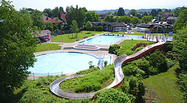 Erlebnisbad Freibad Schwanenstadt in Schwanenstadt: Ein gepflegter, großer Freizeitbereich mit einem Außenpool und einer gewundenen Rutsche. Die Anlage ist von grünen Bäumen und Sträuchern umgeben, die eine ländliche, friedliche Atmosphäre schaffen. Es scheint sich um eine touristische Einrichtung zu handeln, die Besucher zum Entspannen und Genießen einlädt.