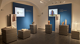 Daringer Kunstmuseum Aspach in Aspach: Ein Ausstellungsraum mit mehreren Kunstwerken und Objekten. Es sind verschiedene Skulpturen und Reliefs zu sehen, die menschliche Figuren und Formen darstellen. Die Werke sind auf Podesten und Regalen platziert und vor einem blauen Wandhintergrund präsentiert. Die Beleuchtung ist hell und fokussiert die Ausstellungsstücke. Insgesamt vermittelt der Raum einen ruhigen und musealen Eindruck.