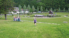 Donau-Spiel Erlebnisspielplatz in Engelhartszell: Eine idyllische Wiese mit einem Brunnen in der Mitte, umgeben von Bäumen und Bergen. Im Vordergrund läuft ein Kind über die Rasenfläche. In der Umgebung sind weitere Menschen zu sehen, die offenbar die Atmosphäre des Ortes genießen.