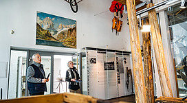 Zwei Personen betrachten Exponate in einem Museum. An der Wand hängt ein Gemälde von Bergen, und verschiedene Ausstellungsstücke sind sichtbar.