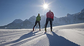 Zwei Skifahrer auf einer verschneiten Piste mit Bergen im Hintergrund. Die Sonne strahlt am klaren Himmel.