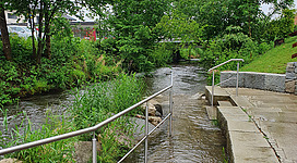 Flößerstrand in Munderfing: Ein altes, steinernes Gebäude mit Grünbewuchs an den Wänden, umgeben von einer idyllischen, bewaldeten Umgebung. Ein Wasserlauf fließt an der Seite des Gebäudes entlang. Verschiedene Gebäudestrukturen und Gitter sind ebenfalls sichtbar.