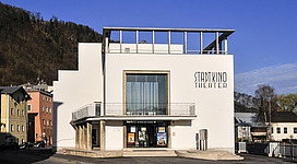 Ein modernes Kino mit einer hellen, weißen Fassade. Vor dem Gebäude sind Werbetafeln für Filme angebracht.