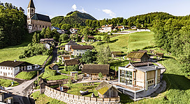 Ein malerisches Dorf inmitten grüner Hügel, umgeben von Bäumen und Bergen. Im Hintergrund ragt eine Kirche mit einem schlanken Turm empor.