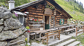 Eine rustikale Berghütte aus Holz mit Blumenschmuck. Draußen stehen Holztische und -bänke, umgeben von einer schönen Landschaft.