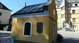 Ein kleines gelbes Gebäude mit einem gewölbten Dach und einem Fenster. Im Hintergrund ist ein modernes Mehrfamilienhaus sichtbar.