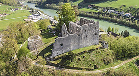 Eine alte Burgruine umgeben von grüner Natur und fließendem Wasser. Im Hintergrund sind sanfte Hügel und Wiesen zu sehen.