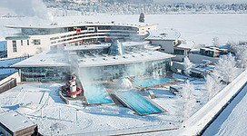 Ein modernes Spa-Hotel in einer schneebedeckten Landschaft. Die großen Fenster zeigen eine elegante Architektur und es gibt einen Außenpool mit dampfendem Wasser.