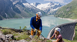 Zwei Kletterer in den Bergen, die an einer Felswand arbeiten. Im Hintergrund sind ein grüner See und schneebedeckte Gipfel zu sehen.