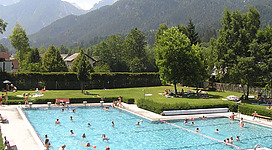 Ein Schwimmbad umgeben von grünen Wiesen und Bergen. Menschen genießen den sonnigen Tag im Wasser und auf Liegen.