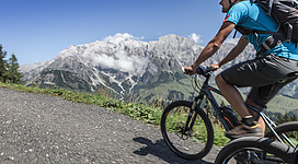 Ein Sportler fährt mit einem Mountainbike auf einem schottrigen Weg. Im Hintergrund sind beeindruckende Berge und ein blauer Himmel mit etwas Wolken zu sehen.